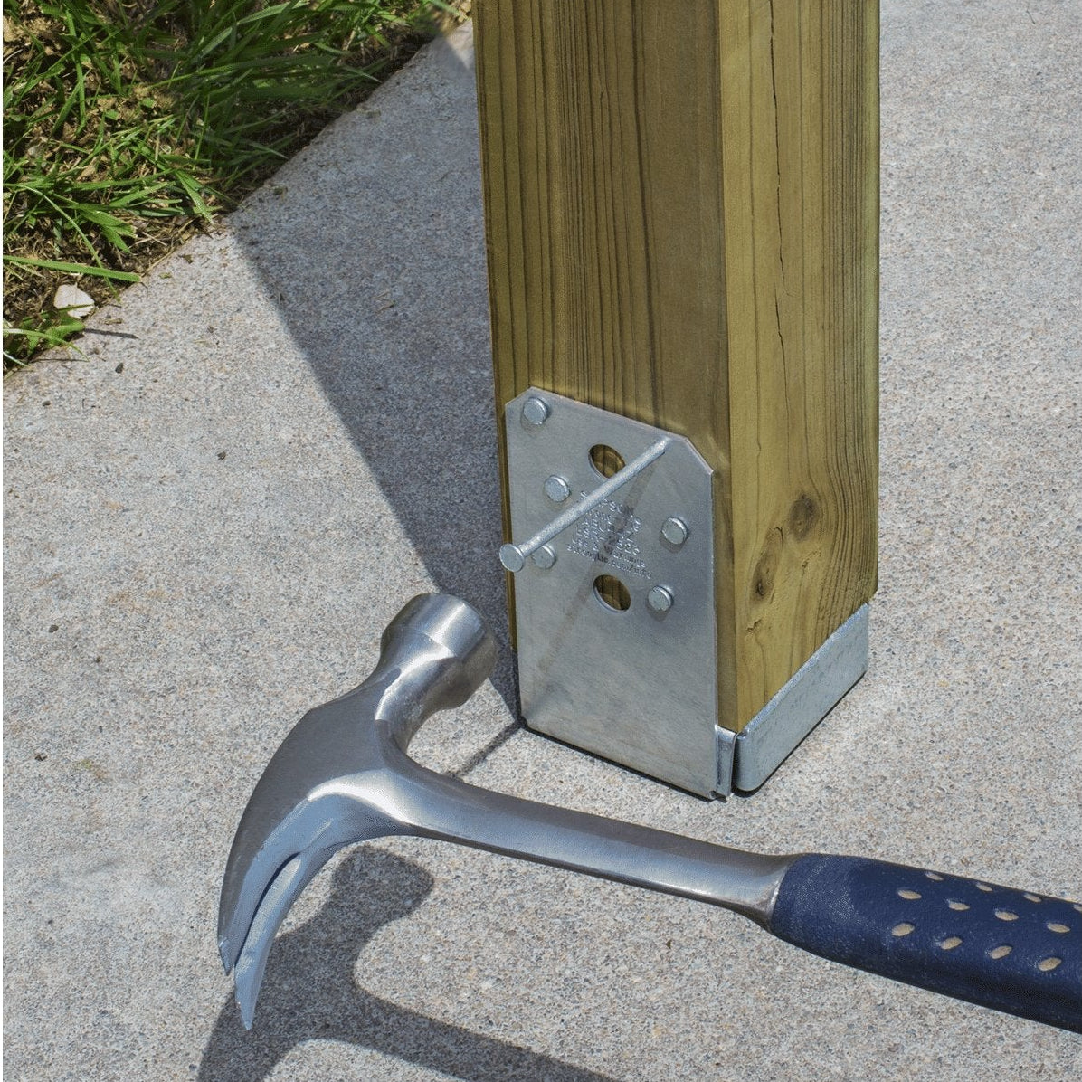 A metal post base anchor with a nail and hammer being installed onto a wooden post on a concrete surface outdoors.