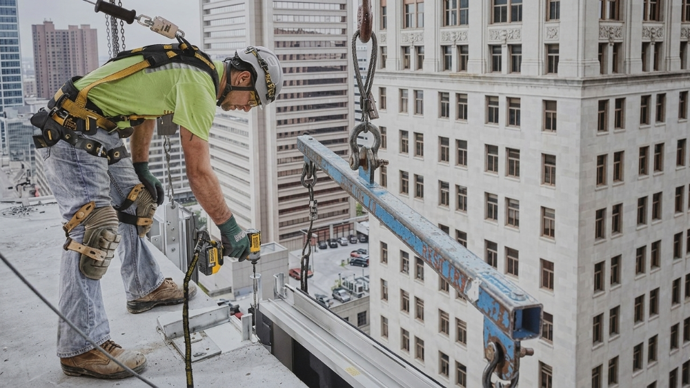 A construction worker in a bright yellow safety shirt and harness stands on a high-rise building rooftop, using a cordless drill near a large blue metal beam suspended by a crane, with city buildings in the background.