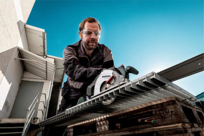 Man using a Metabo circular saw on a construction site with a clear blue sky.