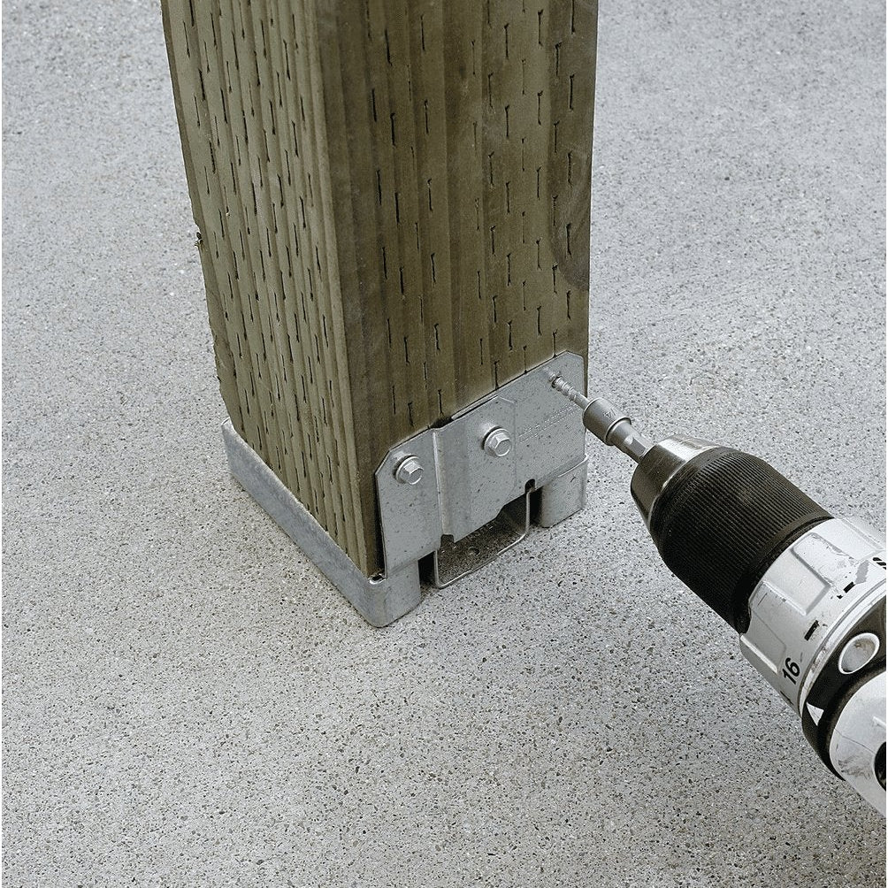 A close-up of a metal post base bracket attached to a wooden post, with a drill screwing a fastener into the bracket. The setup is shown on a light gray concrete surface.