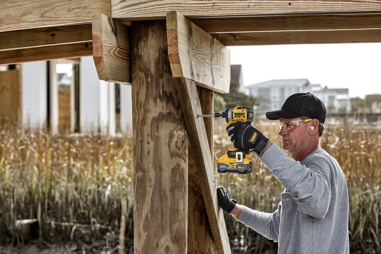 A man wearing safety glasses, a black cap, and gloves uses a yellow and black cordless drill to secure a wooden post in an outdoor construction setting.
