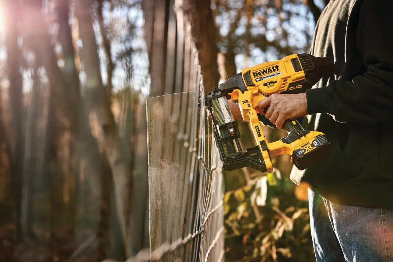 A person using a yellow and black DeWalt cordless nail gun to attach a clear panel to a wooden fence outdoors in warm sunlight.