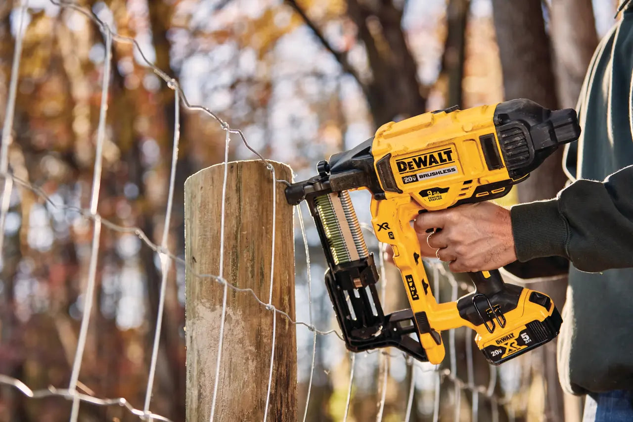 A person using a yellow and black DeWalt cordless nail gun to attach wire to a wooden fence post outdoors, with blurred trees in the background.