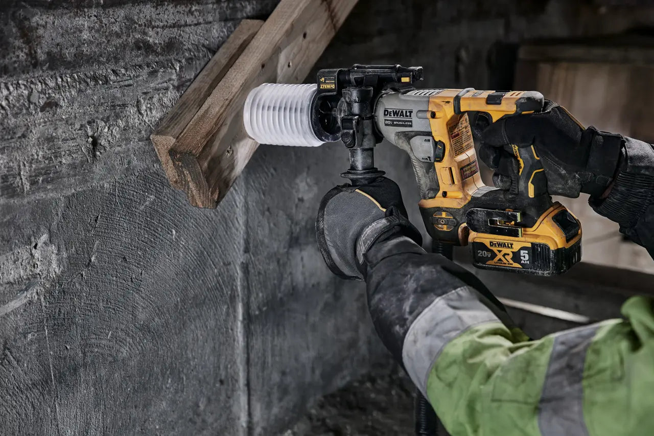 A DeWalt power drill with a white ribbed dust collector attachment being used by a person wearing black gloves and a green high-visibility jacket against a rough concrete wall.
