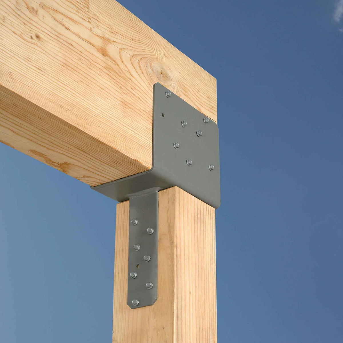 A metal post base bracket installed on a wooden post, with a horizontal beam crossing above, shown against a clear blue sky.
