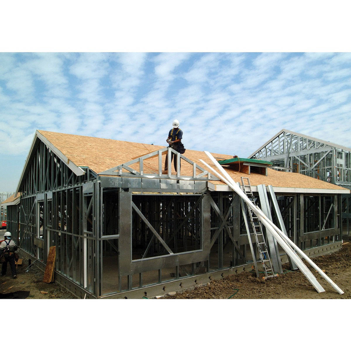 A large metal building frame under construction with a wood-look roof, shown outdoors on a construction site with workers and ladders.