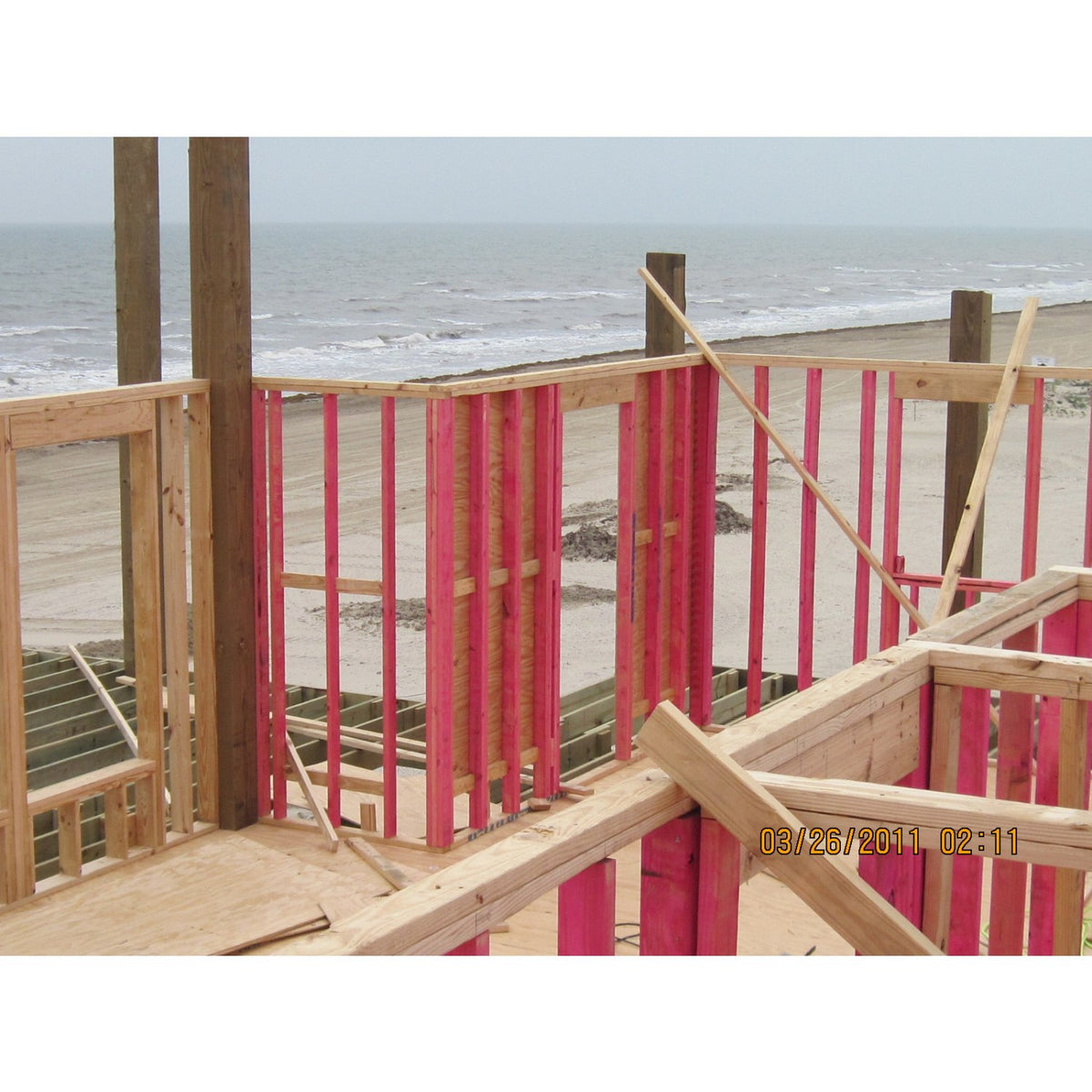A construction scene at the beach showing a wooden deck or structure under frame, with pink formwork panels and natural wood studs. The ocean and sandy shore are visible in the background.