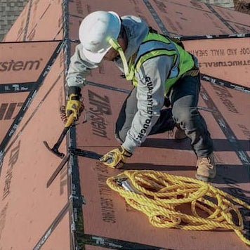 A construction worker wearing a white hard hat, gray jacket, and high-visibility vest is installing or securing yellow rope on a roof. The worker is positioned on top of brown structural decking boards with visible 'System' branding.